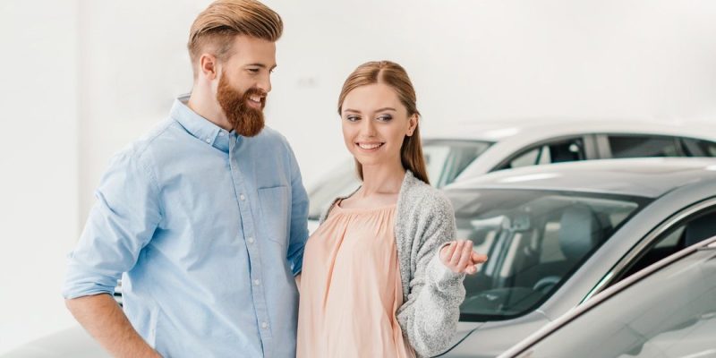 happy-couple-choosing-car-in-dealership-salon-woman-pointing-on-car-e1616394658847.jpg